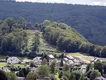 Maisons basses au pied d'une butte boisée surmontée de ruines. Forêt à flanc de coteau à l'arrière-plan.