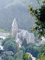 Entourée de maisons basses et d'arbres, l'église à la toiture d'ardoises se détache de la forêt en arrière-plan. Son plan forme une croix flanquée d'un clocher carré.