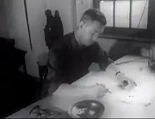 Photographie en noir et blanc d'un homme aux cheveux courts tenant un crayon de la main droite, légèrement penché en avant