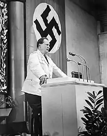 Photographie en noir et blanc de Hermann Göring, debout derrière un pupitre, s'adressant au Reichstag. Derrière lui, une bannière à croix gammée