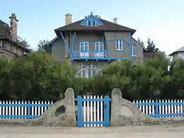 Photo en couleur d'une façade de maison avec fenêtres arrondies et colombages, derrière une palissade et dans la verdure