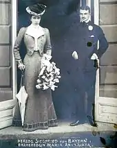 Photo en noir et blanc d'un couple : la fiancée est en longue tenue claire, appuyée sur une ombrelle fermée, aux côtés de son fiancé en costume de ville sombre.