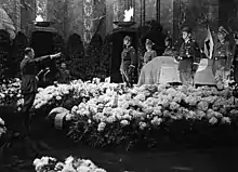 Photographie en noir et blanc de Hitler effectuant le salut nazi devant le cercueil de Heydrich, entouré de fleurs, le 9 juin 1942