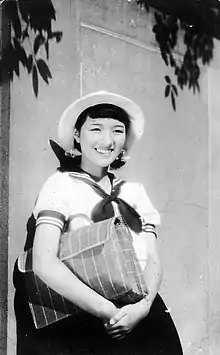 Photographie en noir et blanc d'une adolescente asiatique, en uniforme scolaire de type marin, adossée contre un mur.