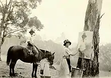 Photographie en noir et blanc. Au premier plan, l'artiste regarde le photographe tout en ayant à la main un pinceau à hauteur de la toile qui est suspendue à un arbre. Au deuxième plan, un homme à cheval de profil. En arrière plan, le bush australine.