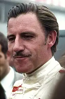 Photographie d'un homme brun, de peau blanche, vu de trois-quarts, avec une moustache, en gros plan, dans une combinaison blanche.