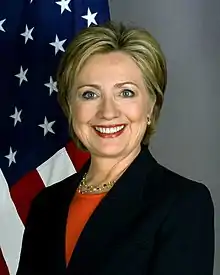 White woman wearing a dark jacket over an orange blouse. The United States flag is in the background.