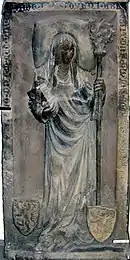 Photographie d'une épitaphe en relief montrant une femme qui tient une crosse et aux pieds de laquelle deux blasons sont posés.