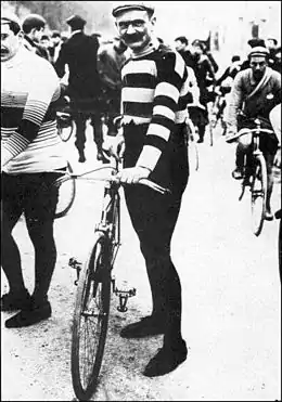 Photographie en noir et blanc d'un homme tenant un vélo et portant un pantalon noir, un haut rayé noir et blanc, de longues moustaches et une casquette.