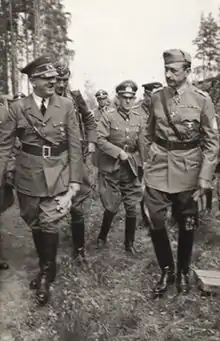 Photo noir et blanc prise le 4 juin 1942. Au premier plan, debout dans l'herbe, Adolf Hitler en uniforme (à gauche) et le maréchal finlandais, Carl Gustaf Emil Mannerheim (à droite), discutent. Au centre, au second plan, un officier allemand, entouré de quelques autres militaires, les suit. Le ciel, en arrière-plan, est clair.