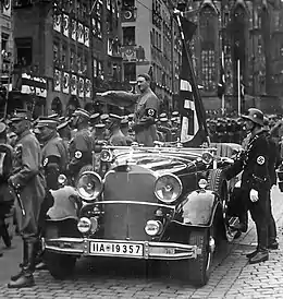 Photographie en noir et blanc de Hitler lors d'un congrès du parti nazi à Nuremberg en 1935. Debout dans une voiture décapotable, il fait le salut nazi à ses partisans qui défilent
