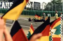 Photo d'une monoplace jaune et verte en piste. Son pilote agite un drapeau allemand.