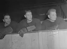 Photographie de 3 joueurs de hockey accoudés derrière la bande de la patinoire.