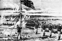 L'Union Jack flotte au sommet d'une colline, hissé par un homme en uniforme militaire. Des officiers et d'autres hommes dans le même uniforme sont au garde-à-vous. Des chariots couverts et des bâtiments de fortune figurent à l'arrière-plan.