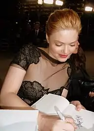 Une jeune femme rousse aux cheveux attachés et en robe de dentelle noire, signe un autographe sur un carnet en souriant légèrement.