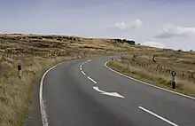 Une lande plate est coupée en deux par une route qui s'éloigne vers la droite sous un ciel nuageux.