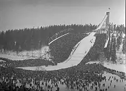 Photographie en noir et blanc d'un tremplin de saut à ski. Une grande foule est présente autour et en bas du tremplin.