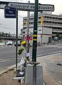 Fleurs et messages au pied d'un panneau et d'un lampadaire ; on voit un panneau portant le nom de la place et une rue en travaux.