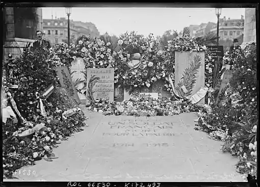 Photo en noir et blanc montrant un amoncellement de couronnes de fleur autour d'une pierre tombale
