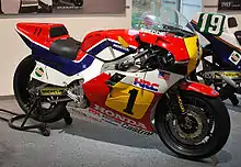 Moto de compétition Honda vue de profil, de couleur rouge, blanche, bleue et jaune.