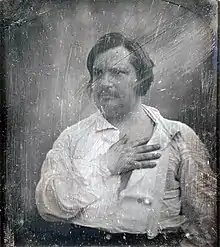 Photographie noir et blanc, Honoré de Balzac, d'âge mûr, cheveux et moustache noirs, vêtu d'une chemise blanche, pose en buste, la main à plat sur le cœur.