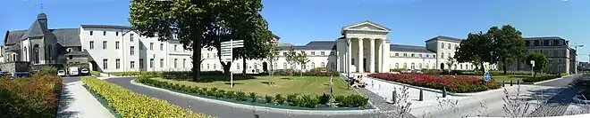 Vue panoramique de l'accès sud de l'hôpital, avec de gauche à droite l'église Saint-Jacques, le pavillon Miséricorde, la chapelle, le pavillon Montfort. Le ciel est bleu, les murs sont blanc, sauf ceux de l'église qui sont gris.