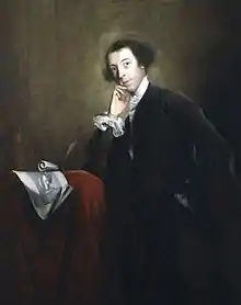 Peinture en clair-obscur d'un homme vêtu de noir ayant le visage éclairé, portant un léger sourire dont l'expression est renforcée par l'index droit posé sur la joue droite.