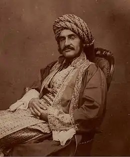 Photographie sépia d'un homme vêtu d'un habit du Moyen-Orient du XIXe siècle, portant une large moustache, assis sur une chaise avec les mains croisées sur ses jambes