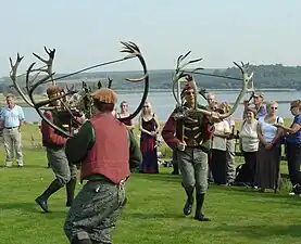 Photo en couleurs de trois hommes vêtus d'habits bariolés qui dansent en rond en portant dans les mains des bois de renne