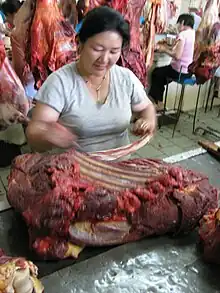 Une femme typiquement asiatique découpe un gros quartier de carcasse sanguinolente le long des côtes à l'aide d'un long couteau, la rapidité de l'action étant suggérée par le flou de la photo.