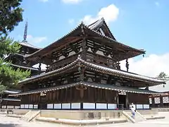 Photo couleur montrant, dressé sur une dalle en pierre beige, un bâtiment en bois à deux étages d'un temple bouddhique (couleurs dominantes de la structure : marron foncé et blanc). À l'arrière-plan, à gauche, une pagode à cinq étages, en bois marron, sur fond de ciel bleu parsemé de quelques nuages.