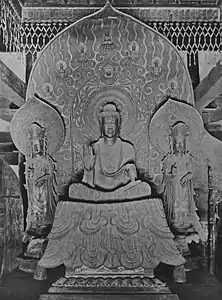 Photo noir et blanc d'une statue en bronze représentant un bouddha en position du lotus, entouré de deux assistants debout.