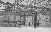 Photo noir et blanc de l'usine en travaux avec de nombreux ouvriers au travail sous la charpente métallique qui n'a pas encore sa toiture