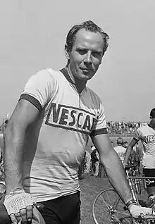 Photographie en noir et blanc montrant un cycliste se tenant debout à côté de son vélo, portant un maillot avec l'inscription Nescafé.