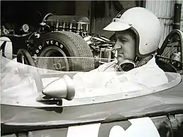 Photographie en noir et blanc du cockpit d'une Formule 1, de profil, avec le pilote avec un casque blanc, dans sa Formule 1.