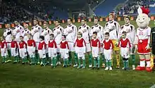 Sur un terrain de football, onze joueurs chantent leur hymne national, la main sur le cœur devant onze enfants.