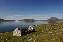 Une église de pierre en ruines, sans toiture, domine une prairie rocailleuse descendant vers un fjord.