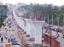 Photographie d'un axe routier important, en Inde, au centre duquel le terre-plein central est remplacé par une rangée de dizaines de hauts poteaux de bétons, en attente de la plate-forme du métro