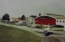 Photographie ancienne représentant quatre avions devant un hangar à la porte rouge. De gauche à droite :  un B-17, un Hurel, un AirCommander et un Mystère 20.