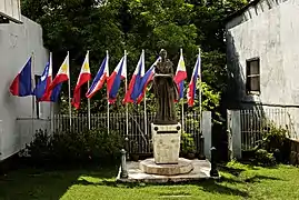 Statue jeune femme debout, vêtu d'une longue robe. Porte un drapeau plié dans ses mains, paumes tournées vers le ciel.