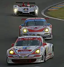 Photographie de trois voitures (une blanche et rouge, deux grise et rouge) de face, sur une piste.
