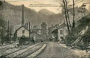 Carte postale ancienne montrant l'entrée des Forges d'Allevard encadrées de montagnes et l'arrivée de la voie à l'écartement de 1,10 m