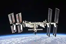 La station est composée de nombreux modules associés à des panneaux solaires, la Terre se trouve en bas de l'image.