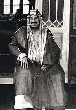 Un homme assis, souriant et moustachu porte une dishdasha sombre et un keffieh.