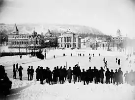 Photo noir et blanc d’un match de hockey joué en extérieur devant une ville enneigée.
