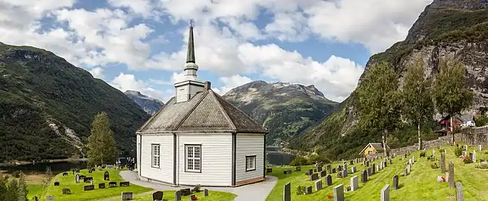 Un petit bâtiment blanc dans les montagnes comportant un unique res-de-chaussée surmonté d'un petit clocher entouré d'une pelouse où s'alignent des pierres tombales.