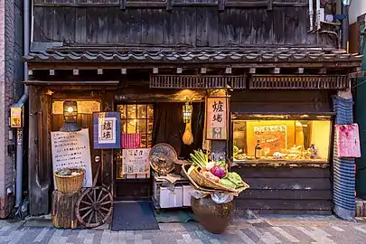 Façade illuminée d'un restaurant servant des plats traditionnels et présentant des paniers de légumes frais à l'entrée, Yūrakuchō. Juin 2019.