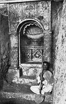 Photo en noir et blanc. Un homme assis tenant une pelle est juste à côté d'une niche.