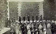 Photomontage en noir et blanc représentant l'exécution de six religieux, debout devant le mur de la cour intérieure d'une prison. Les religieux sont tenus en joue par un peloton au premier plan, qu'un homme sur le côté gauche semble commander.