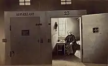 Photomontage en noir et blanc présentant, en intérieur, deux portes de prison, dont celle de droite ouverte laisse voir un religieux assis dans sa cellule.